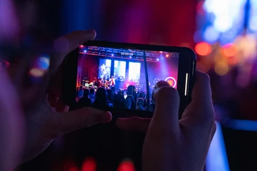 Stadthalle Bremerhaven_HAI_DANG_DUONG_00271.jpg Aufnahme eines Smartphones, das eine Konzertbühne mit bunter Beleuchtung filmt.