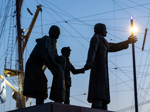 Dunkle Bronzestatue einer Auswandererfamilie vor beleuchteten Schiffsmasten in der Dämmerung.