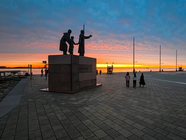 IMG_20241008_190311_418 (c) Helmut Gross_Erlebnis Bremerhaven.jpg Bronzeplastik einer Auswandererfamilie auf Steinsockel am Hafenbecken bei Sonnenuntergang.