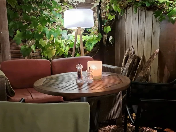 Hafen5_4.png Abendliche Terrasse mit beleuchtetem Holztisch, Kerze, weichen Sesseln und grünen Weinranken.