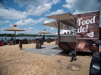 Foodtruck 1200_Gastro_Beach.jpg Foodtruck an Strandbar mit Palmen-Sonnenschirmen, Sitzfässern und Seeblick bei sonnigem Wetter.