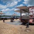Foodtruck 1200_Gastro_Beach.jpg Foodtruck an Strandbar mit Palmen-Sonnenschirmen, Sitzfässern und Seeblick bei sonnigem Wetter.
