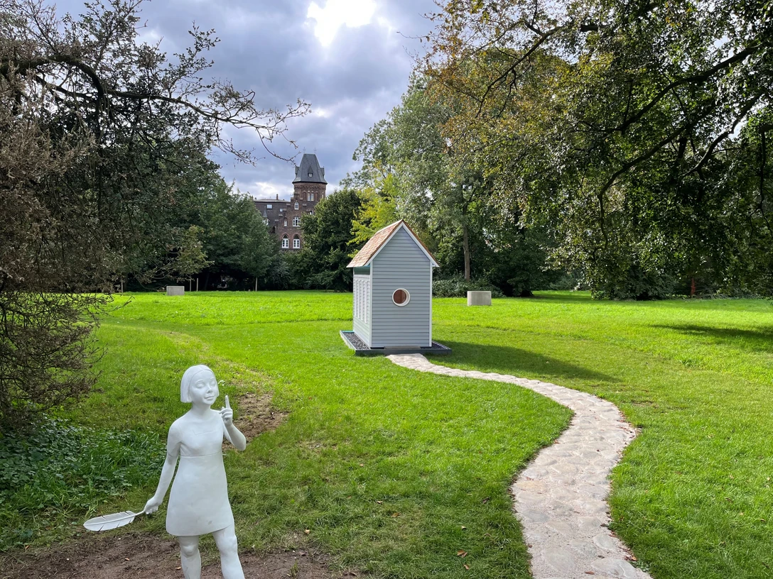 Monheim am Rhein künstlerisch entdecken Kunstinstallation mit Mädchenstatue, kleinem Haus, Steinweg und historischem Gebäude in Monheim am Rhein.
