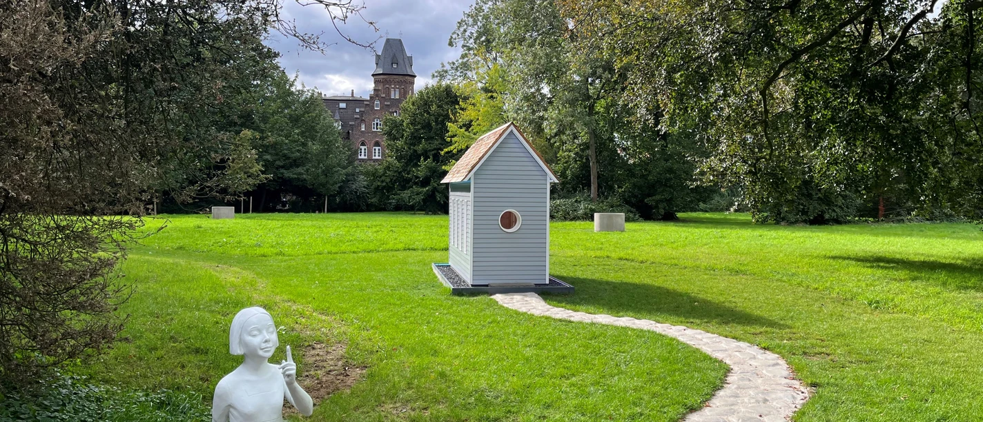 Monheim am Rhein künstlerisch entdecken Kunstinstallation mit Mädchenstatue, kleinem Haus, Steinweg und historischem Gebäude in Monheim am Rhein.