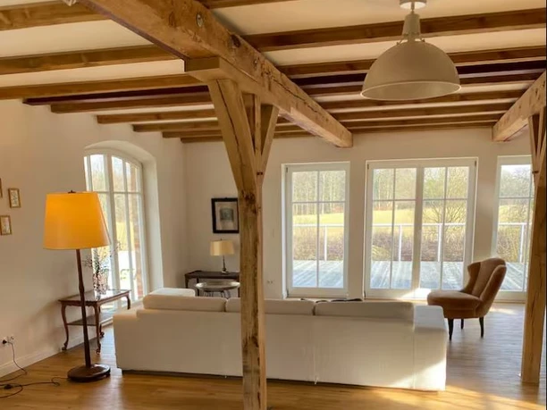 modernes-wohnzimmer-fachwerk-samtgemeinde.jpg.JPG Ein helles Wohnzimmer mit moderner Couch, rustikalen Holzbalken und großen Fenstern mit Blick ins Grüne.