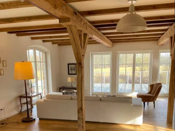 modernes-wohnzimmer-fachwerk-samtgemeinde Ein helles Wohnzimmer mit moderner Couch, rustikalen Holzbalken und großen Fenstern mit Blick ins Grüne.