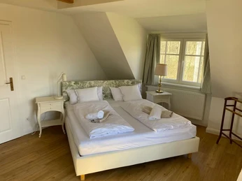 helles-schlafzimmer-dachgeschoss-immobilie.jpg.JPG Ein gemütliches Doppelbett unter einer Dachschräge mit hellen Möbeln und Blick aus dem Fenster.