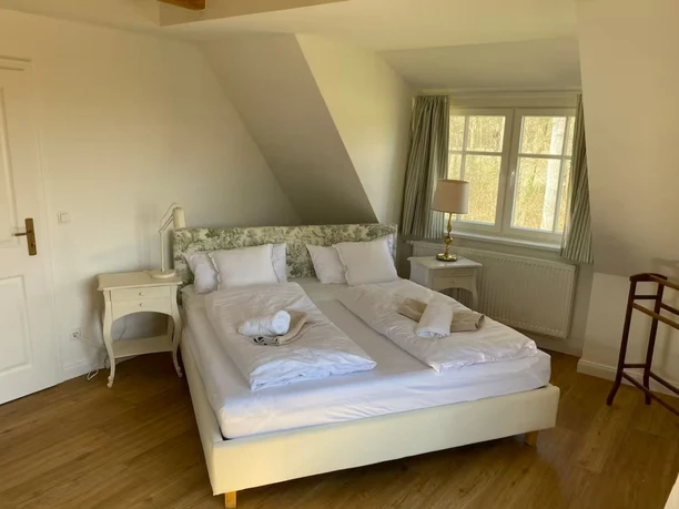 helles-schlafzimmer-dachgeschoss-immobilie Ein gemütliches Doppelbett unter einer Dachschräge mit hellen Möbeln und Blick aus dem Fenster.