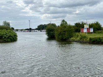 Weyhe-Dreye Wieltsee Einfahrt Wasserweg zum Wieltsee mit grünen Ufern, Booten im Hafen und Windrädern im Hintergrund.