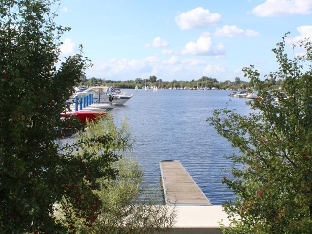 Wieltsee Holzsteg am Wieltsee, umrahmt von grünen Büschen. Auf dem Wasser liegen mehrere weiße Boote.