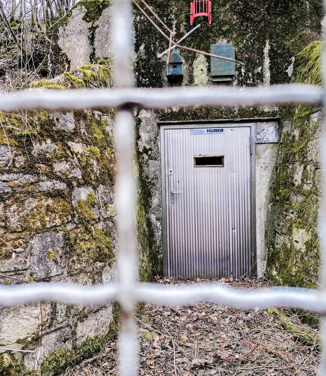 Hochbehälter 2.jpg Blick durch einen Maschendrahtzaun auf eine Metalltür eines von Moos bewachsenen Steingebäudes.