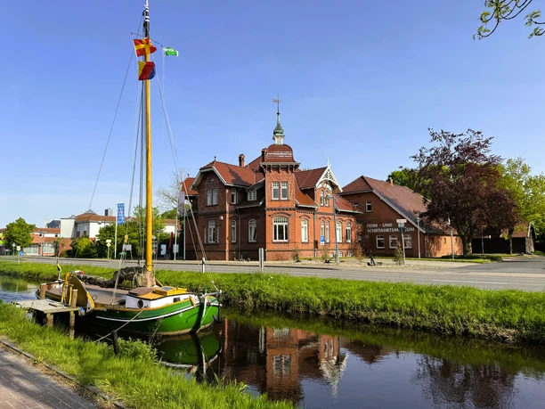 Außenbereich der Tourist-Information Rhauderfehn Grünes Segelboot am Ufer eines Kanals vor einem roten Backsteinhaus mit Ziegeldach und Türmchen bei klarem Himmel