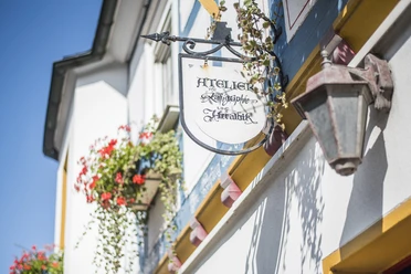 Atelier für Heraldik, Hatzfeld (Eder) Sie sehen ein kunstvoll gestaltetes, schmiedeeisernes Werbeschild an der Fassade, das in eleganter Schreibschrift auf das „Atelier für Kalligrafie & Heraldik“ hinweist.