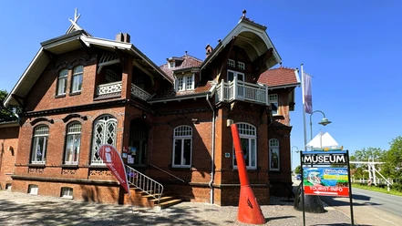 Eingang_Tourist_Info_Rhauderfehn_Juni_2024_S.K..png Backsteingebäude mit verziertem Dach und mehreren Fenstern neben einem Schild mit der Aufschrift 'MUSEUM' und 'TOURIST-INFO'