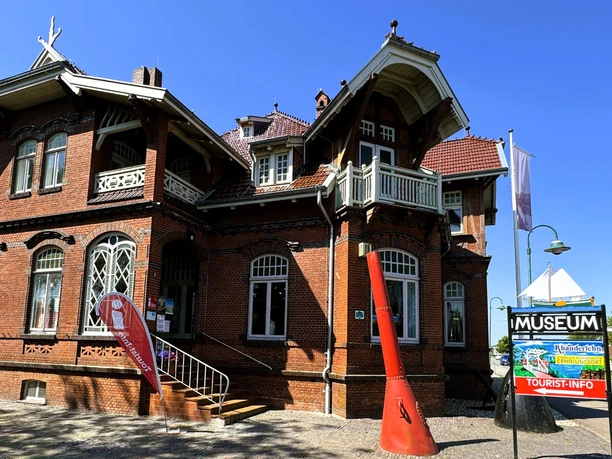 Eingang_Tourist_Info_Rhauderfehn_Juni_2024_S.K..png Backsteingebäude mit verziertem Dach und mehreren Fenstern neben einem Schild mit der Aufschrift 'MUSEUM' und 'TOURIST-INFO'