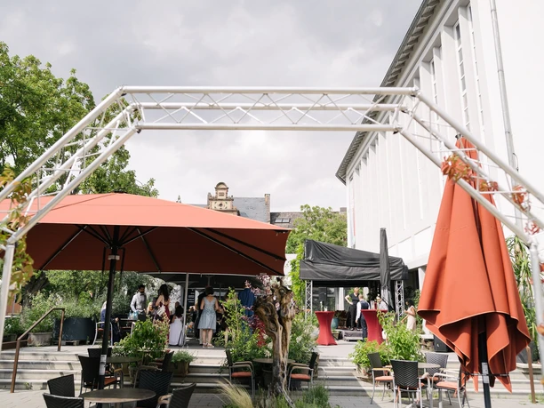 RheinMain Hanau AJOKI - Terrasse Außenbereich eines Cafés mit Gästen, Sonnenschirmen und Bühne