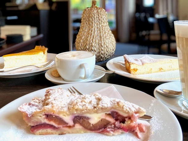 Kaffee und Kuchen im Café Grunewald En Stück Pflaumenkuchen liegt auf einem Teller, dahinter ist verschwommen das Café zu sehen.