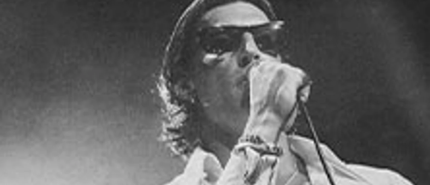 Sänger mit Sonnenbrille und Mikrofon auf einer dunkel ausgeleuchteten Bühne.