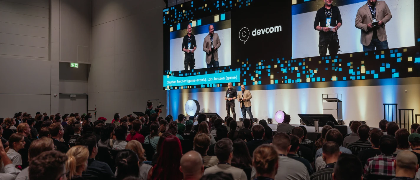 Gamescom dev People sit in well-filled rows facing the stage, on which two presenters are standing. The screen in the background shows the words "devcom".