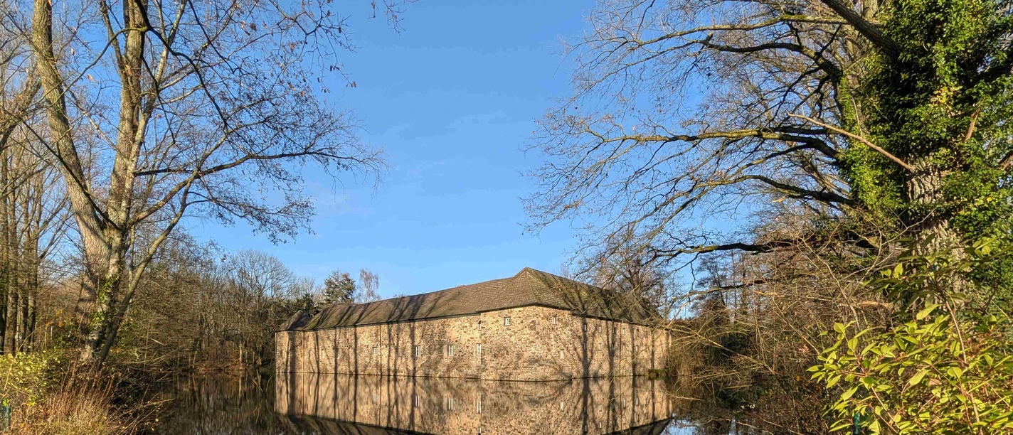 Gesundheitswanderung Steinernes Gebäude am Wasser mit kahlen Bäumen und deren Spiegelung unter blauem Himmel.