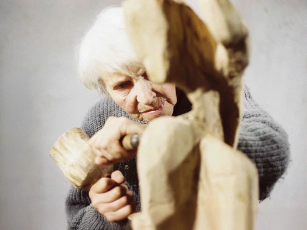 Louise Stomps at work on her last sculpture "Der Aussteiger", Rechtmehring 1987 Elderly person working intently with a mallet and chisel on a rough wooden sculpture.