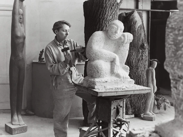 Elsa Thiemann (1910-1981), Louise Stomps in the studio at work on "Sitzenden", Berlin 1946/47, repro print, Berlinische Galerie Person works on a large stone figure in a studio with other sculptures.