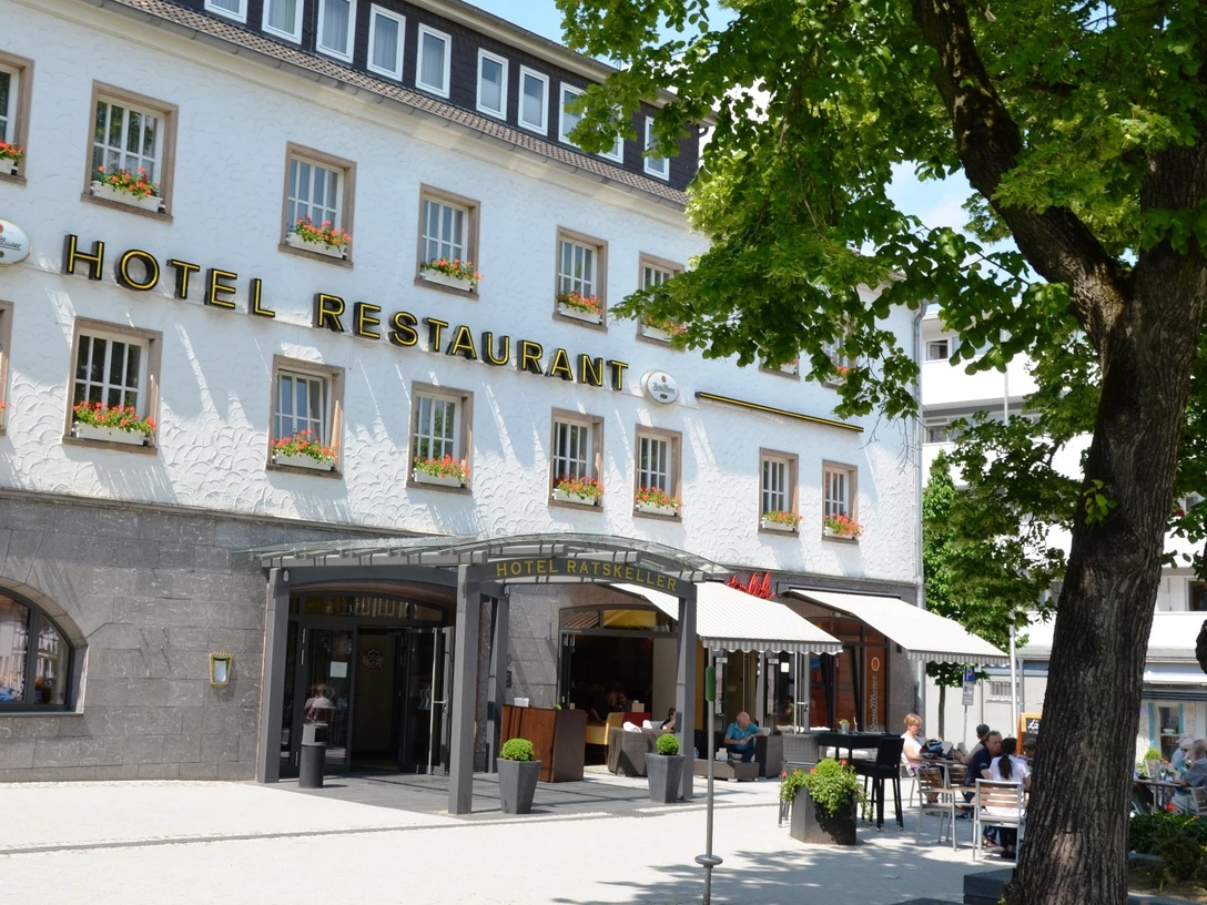 Weißes Gebäude des Hotels Ratskeller mit Außengastronomie unter Bäumen.
