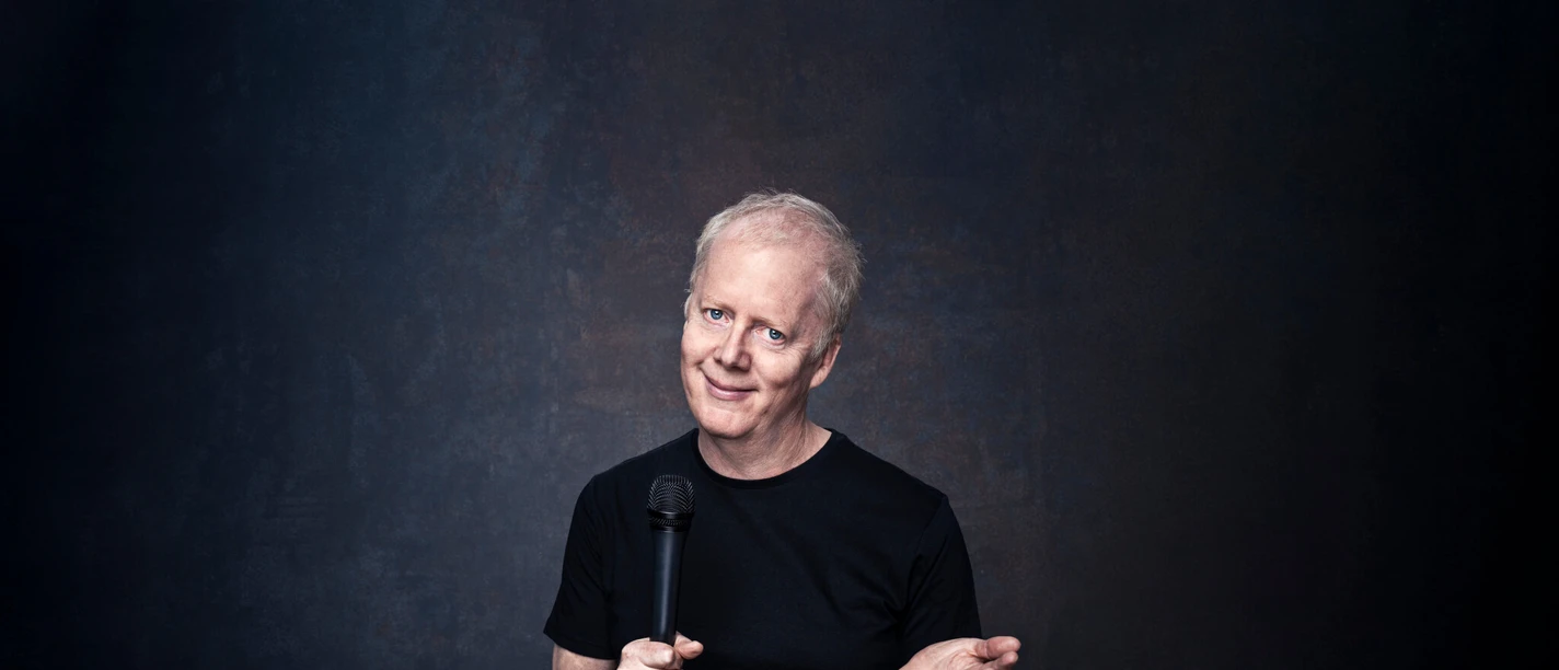 John Doyle Man with microphone in front of dark background with slight smile and open gesture