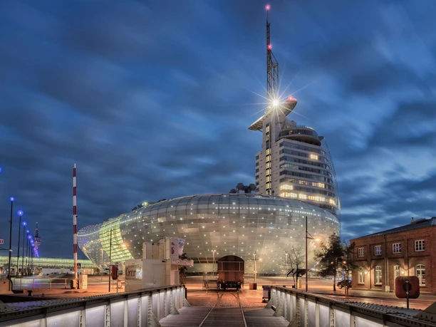Klimahaus Bremerhaven Außenansicht 2 3000px(c) Thorsten Ernst_WEB.jpg Blick über eine Brücke mit dahinter stehendem Bahnwaggon auf das beleuchtete Klimahaus Bremerhaven