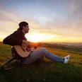 Mattis Brodersen in Büsum Ein Mann mit Cap sitzt mit Gitarre auf dem Deich. Im Hintergrund geht die Sonne unter.