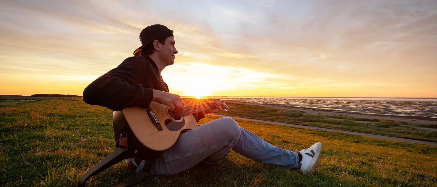 Mattis Brodersen in Büsum Ein Mann mit Cap sitzt mit Gitarre auf dem Deich. Im Hintergrund geht die Sonne unter.