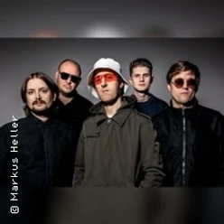 Fünf Männer in Studioatmosphäre, der mittlere trägt weißen Hut und rote Brille.