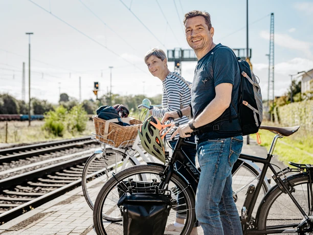 Met de fiets naar het station Twee fietsers staan met hun fiets bij een treinstation, naast het spoor, klaar om hun reis met de trein voort te zetten.