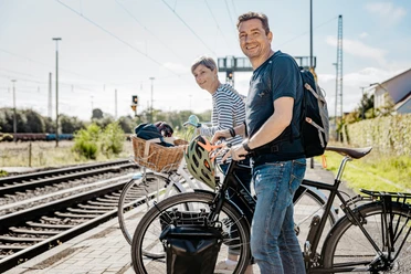 Mit dem Rad am Bahnhof Zwei Radfahrende stehen mit ihren Fahrrädern an einem Bahnhof, neben den Gleisen, bereit für die Weiterreise mit der Bahn.