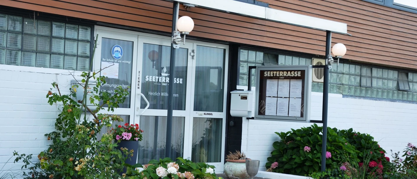 Eingangsbereich des Restaurants Seeterrasse mit Glastüren, Speisekartenkasten und Blumenbeeten.