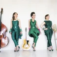Zucchini Sistaz Drei Frauen in grünen Outfits stehen mit Kontrabass, Gitarre und Posaune vor einem weißen Hintergrund.