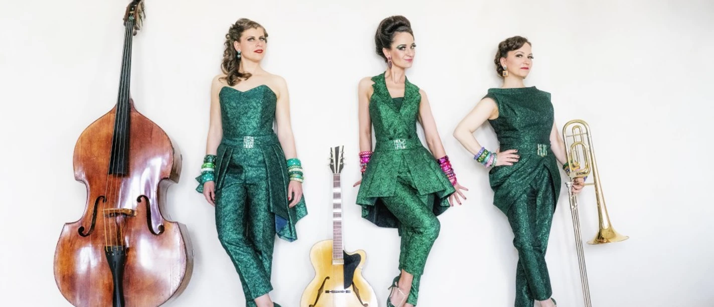 Zucchini Sistaz Drei Frauen in grünen Outfits stehen mit Kontrabass, Gitarre und Posaune vor einem weißen Hintergrund.