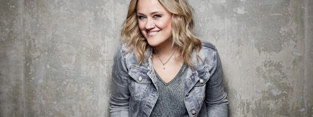 A woman smiling in a gray denim jacket in front of a gray concrete wall.