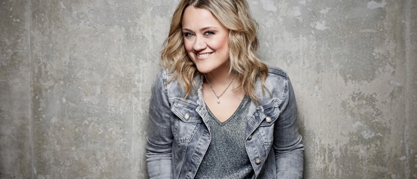 A woman smiling in a gray denim jacket in front of a gray concrete wall.
