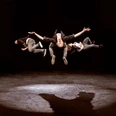 Three men in a leaping position on stage, on a dark background.