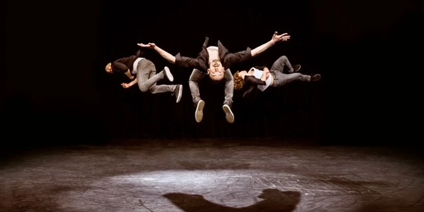 Three men in a leaping position on stage, on a dark background.