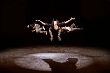 Tridiculous Drei Männer in Luftsprungposition auf der Bühne, auf dunklem Hintergrund.