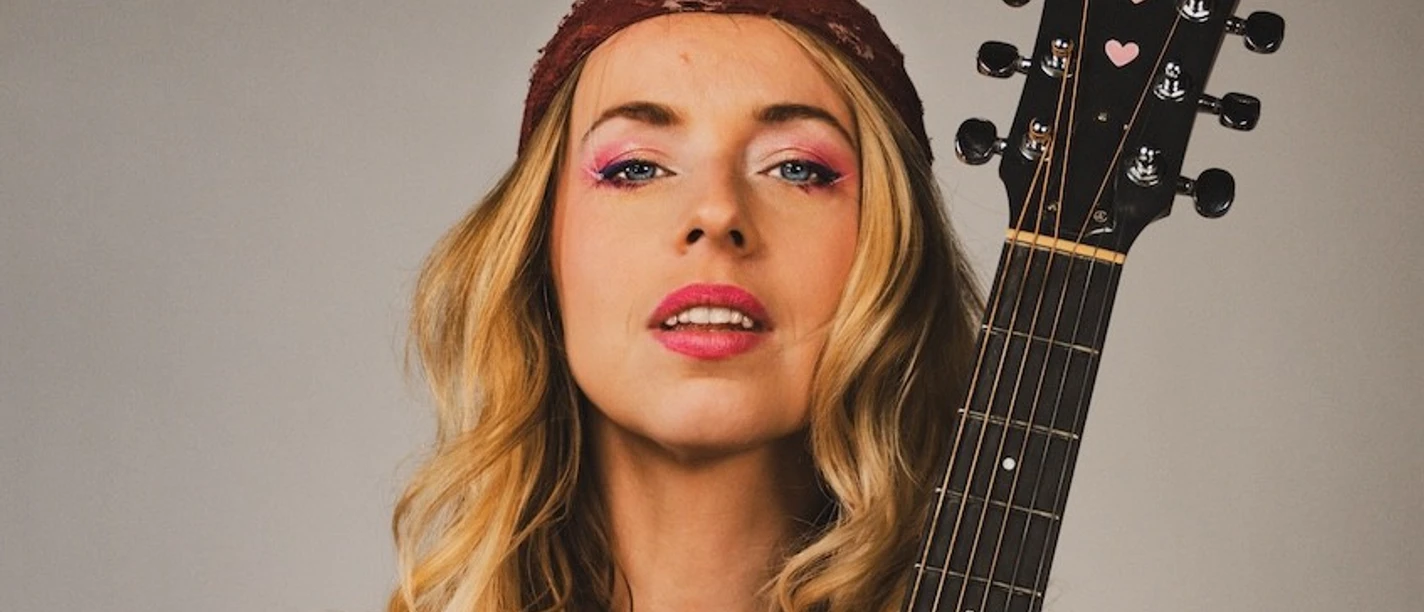 Miss Allie Frau mit Gitarre in der Hand, trägt weißes Oberteil und rotes Stirnband, vor grauem Hintergrund.