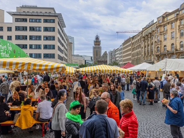 Grüne Soße Festival Menschenmenge auf einem belebten Marktplatz mit gelb-weiß gestreiften Zelten und Gebäuden im Hintergrund.