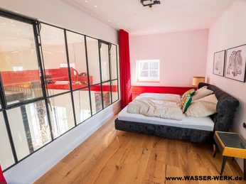 Schlafzimmer Pumpensaal.jpg Schlafzimmer mit Doppelbett, Glaswand zum Pumpensaal, Holzboden und roten Vorhängen