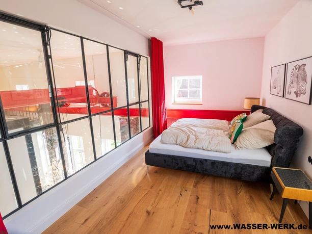 Schlafzimmer Pumpensaal.jpg Schlafzimmer mit Doppelbett, Glaswand zum Pumpensaal, Holzboden und roten Vorhängen