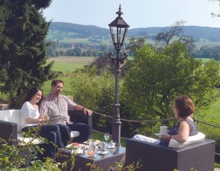 Auf der Sonnenterasse Drei Gäste sitzen auf einer Terrasse mit Blick über grüne Hügel und trinken Kaffee.