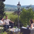 Auf der Sonnenterasse Drei Gäste sitzen auf einer Terrasse mit Blick über grüne Hügel und trinken Kaffee.