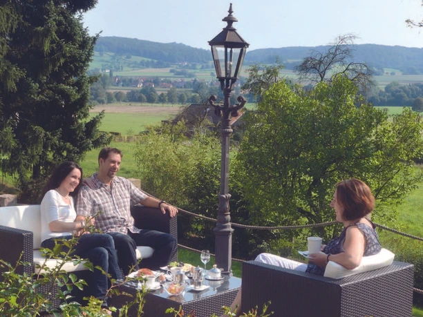 Auf der Sonnenterasse Drei Gäste sitzen auf einer Terrasse mit Blick über grüne Hügel und trinken Kaffee.