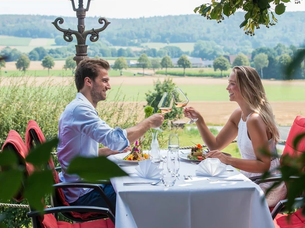 Essen mit herrlichem Ausblick Paar stößt auf Terrasse mit Wein an, gedeckter Tisch, Blick über Felder.